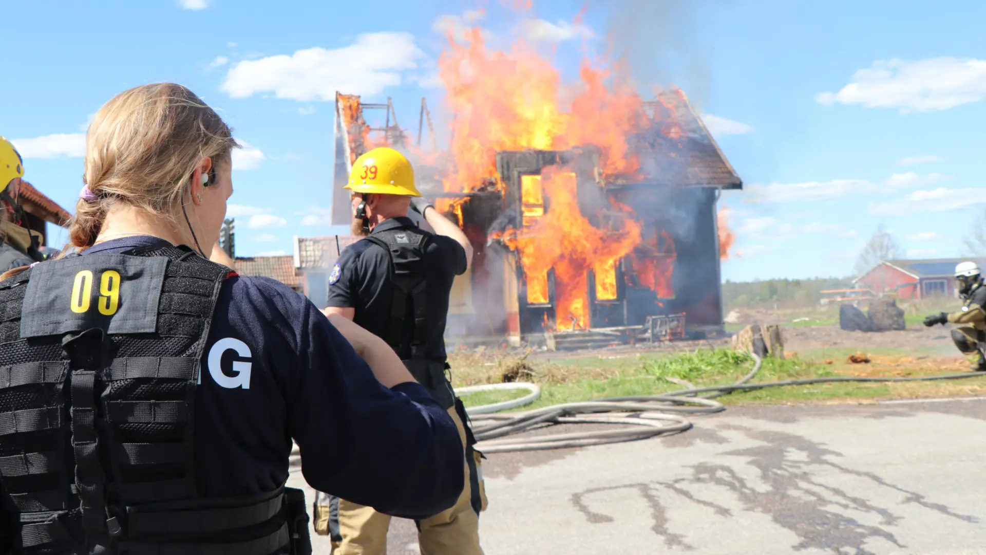 Ett hus som brinner i bakgrunden, I förgrunden två brandmän